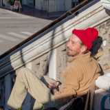 Quando lo incontriamo � seduto sui gradini, indossa un cappello rosso sui capelli biondi ed � intento a parlare con un passante